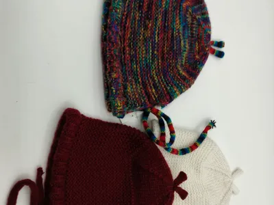 Drei handgestrickte Babyhäubchen mit Kinnbändern nebeneinander in buntem Farbverlaufsgarn, satten Burgunderrot und klassischem Cremeweiß, auf weißem Untergrund