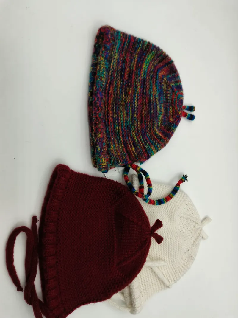 Drei handgestrickte Babyhäubchen mit Kinnbändern nebeneinander in buntem Farbverlaufsgarn, satten Burgunderrot und klassischem Cremeweiß, auf weißem Untergrund