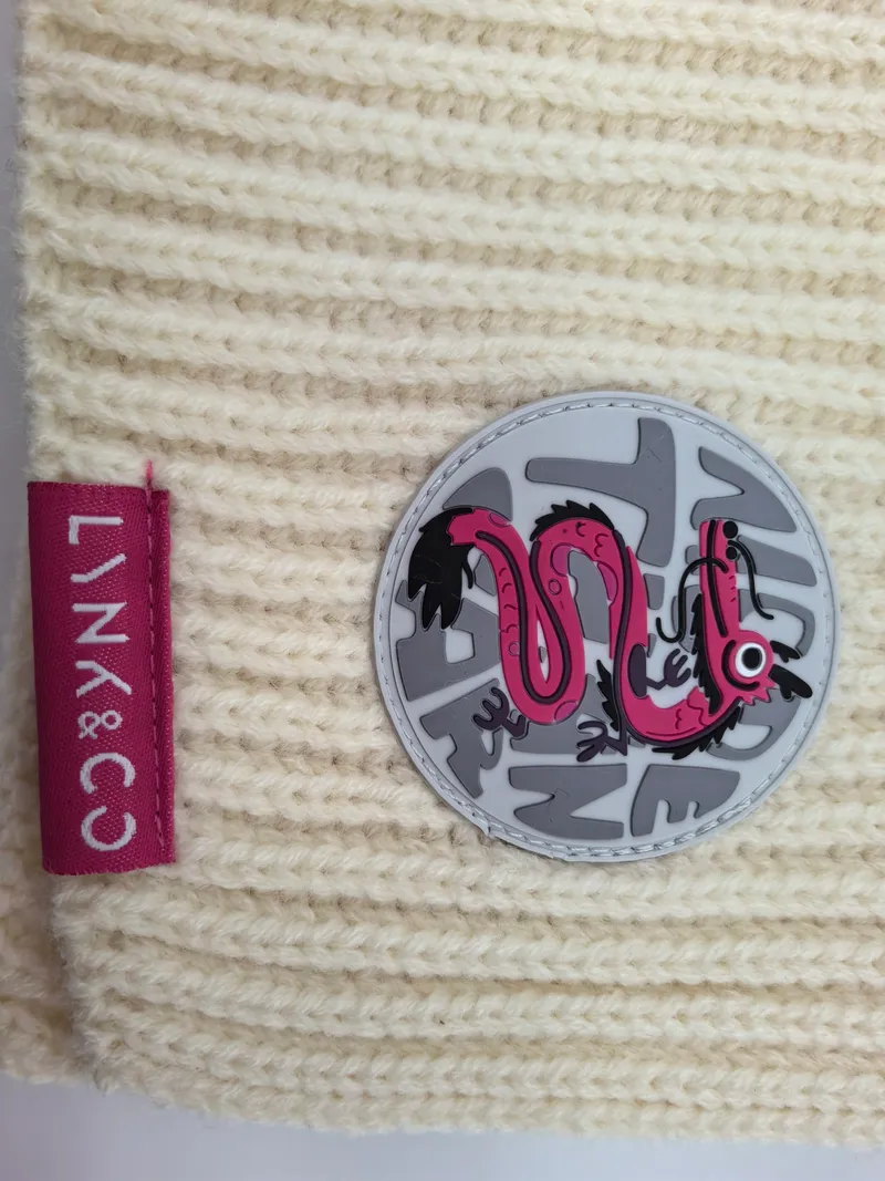 Hellbeige Wintermütze in grobem Rippenstrick mit rundem rosa Gummiabzeichen und Drachenmotiv sowie farbigem Markenlabel auf strukturierter Oberfläche