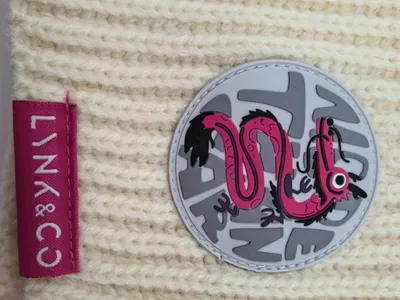 Hellbeige Wintermütze in grobem Rippenstrick mit rundem rosa Gummiabzeichen und Drachenmotiv sowie farbigem Markenlabel auf strukturierter Oberfläche