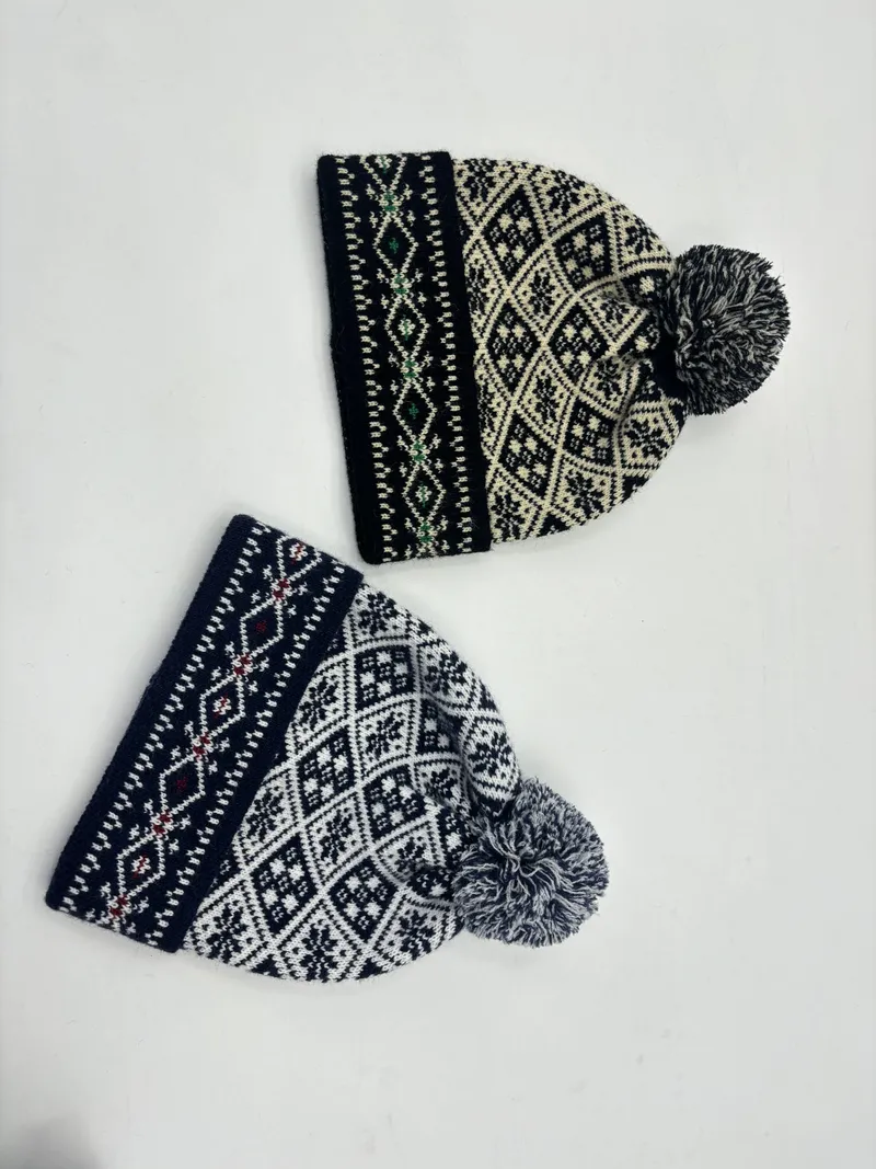 Zwei Strickbommelmützen im skandinavischen Fair-Isle-Muster auf weißem Grund: eine in Schwarz-Creme, eine in Marineblau-Weiß mit Schneeflocken-Rautenmotiv