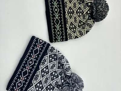 Zwei Strickbommelmützen im skandinavischen Fair-Isle-Muster auf weißem Grund: eine in Schwarz-Creme, eine in Marineblau-Weiß mit Schneeflocken-Rautenmotiv
