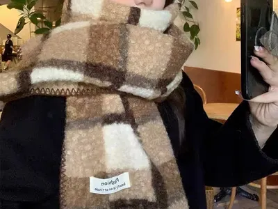 Junge Frau trägt einen dicken, flauschigen Karoschal in Beige-Braun-Creme-Tönen und macht drinnen ein Selfie