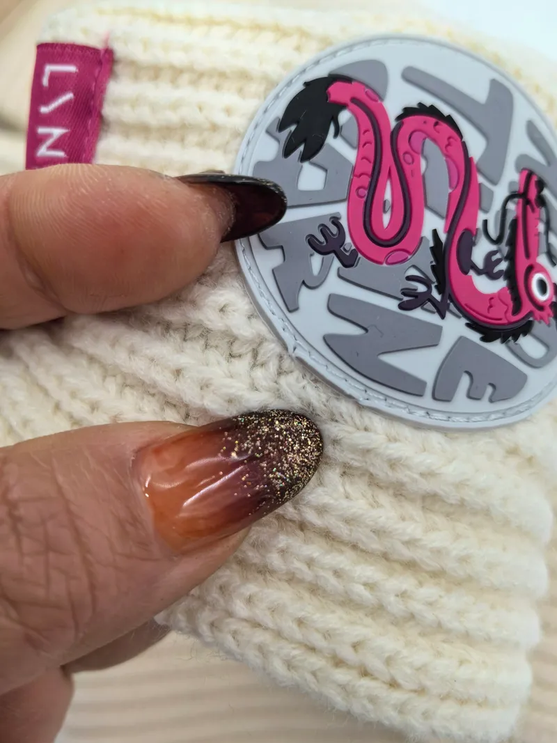 Nahaufnahme einer cremeweißen Wintermütze in der Hand mit glitzernden Ombré-Nägeln, verziert mit rundem rosafarbenem Gummipatch mit Schlangenmotiv