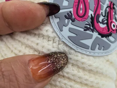 Nahaufnahme einer cremeweißen Wintermütze in der Hand mit glitzernden Ombré-Nägeln, verziert mit rundem rosafarbenem Gummipatch mit Schlangenmotiv
