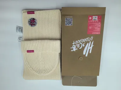 Cremefarbene Rippenstrickmütze und passender Schal mit burgunderrotem Logoaufnäher, präsentiert neben einer Kraftpapier-Geschenkbox