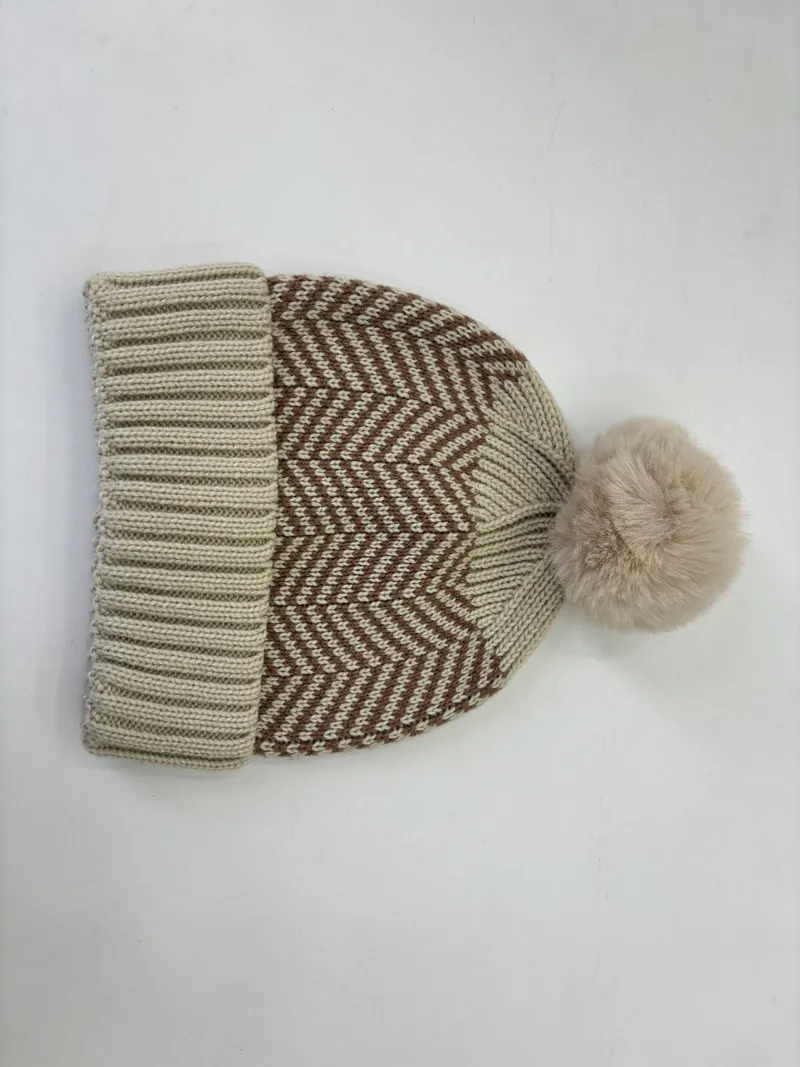 Beige-braune Wintermütze mit Chevron-Strickmuster und großem flauschigem Pompom aus Kunstfell vor weißem Hintergrund