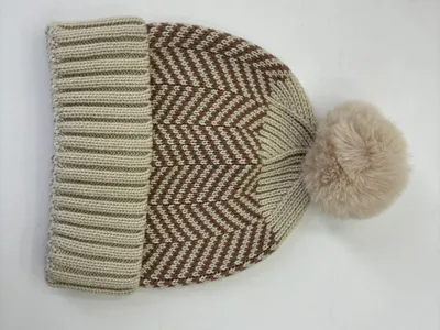 Beige-braune Wintermütze mit Chevron-Strickmuster und großem flauschigem Pompom aus Kunstfell vor weißem Hintergrund