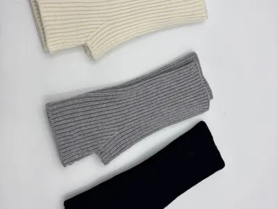 Drei fingerlose Rippenstrick-Armstulpen nebeneinander in den Farben Elfenbein, Hellgrau und Schwarz, flach auf weißem Hintergrund