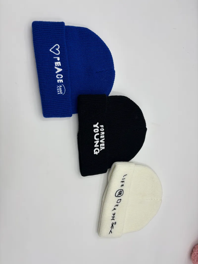 Drei gerippte Umschlagmützen in Blau, Schwarz und Creme mit bestickten englischsprachigen Slogans Peace, Forever Young und Life or Death