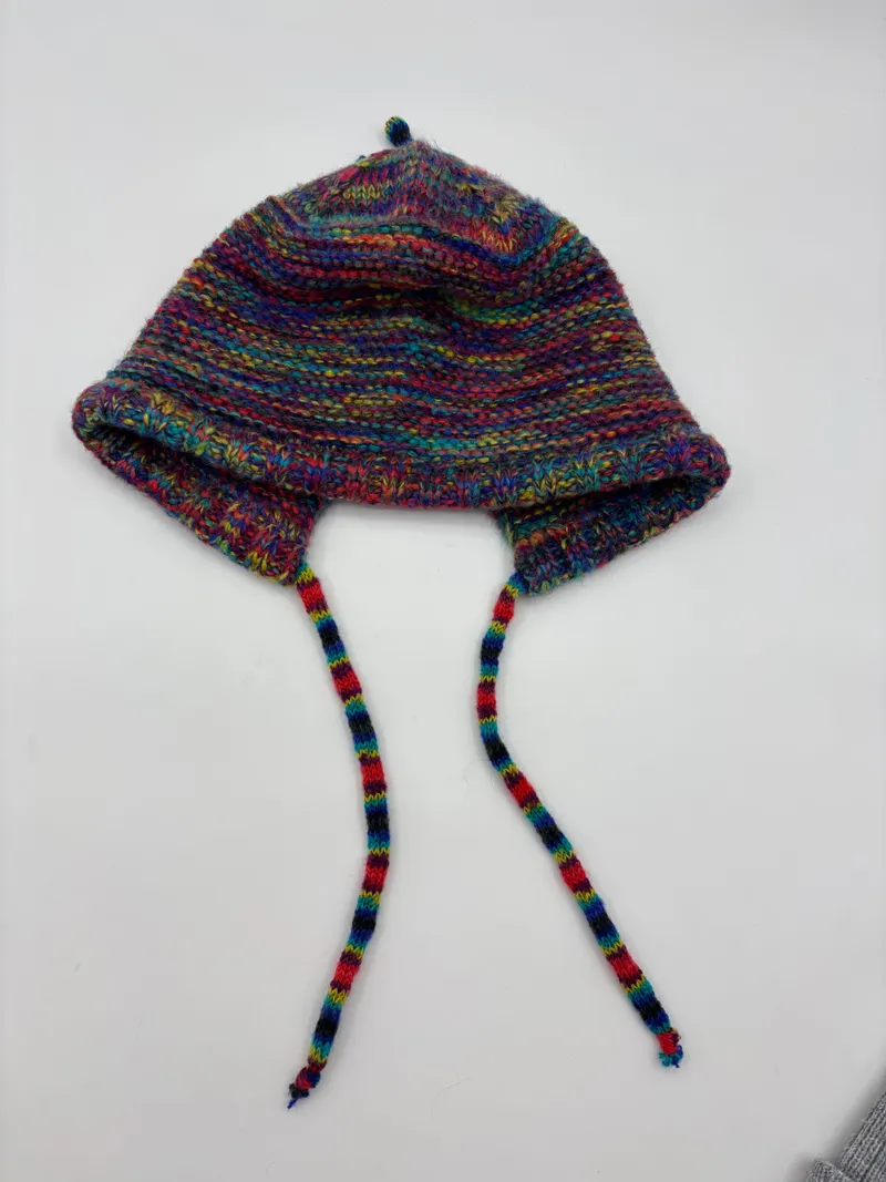 Handgestrickte Babyhäubchen aus mehrfarbigem Buntgarn mit Ohrenschutz und geflochtenen Bindebändern, Flachlage auf weißem Grund