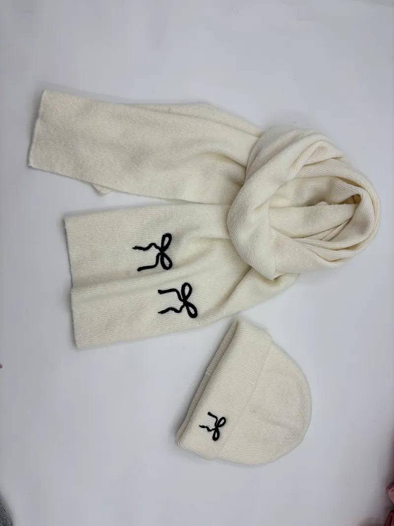 Cremeweißes Rippenstrick-Winterset aus Mütze und Schal mit bestickten schwarzen Schleifenmotiven, flach auf hellem Untergrund