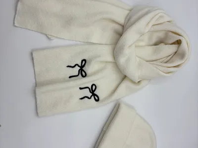 Cremeweißes Rippenstrick-Winterset aus Mütze und Schal mit bestickten schwarzen Schleifenmotiven, flach auf hellem Untergrund