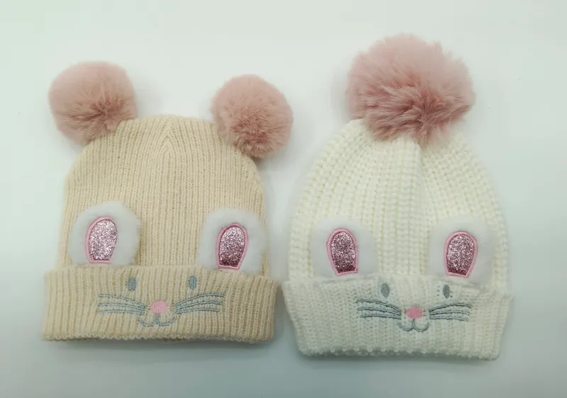Zwei gestrickte Baby-Mützen in Creme und Weiß mit glitzernden Hasenohren, aufgestickten Schnurrhaaren und weichen pinken Kunstfell-Pompons oben
