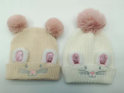 Zwei gestrickte Baby-Mützen in Creme und Weiß mit glitzernden Hasenohren, aufgestickten Schnurrhaaren und weichen pinken Kunstfell-Pompons oben
