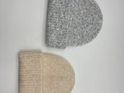 Zwei Rippenstrickmützen mit Umschlagbündchen aus Wollmischung – eine in Heathergrau, eine in Cremeweiß, auf weißem Hintergrund