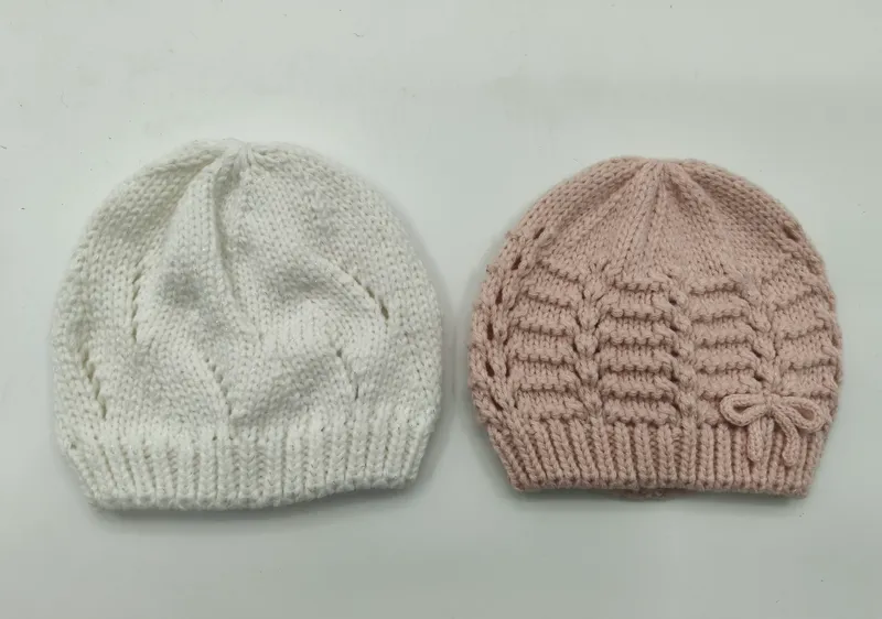 Zwei handgestrickte Babymützen nebeneinander, eine weiß mit Blattmuster und eine altrosa mit horizontaler Textur und Schleifendetail
