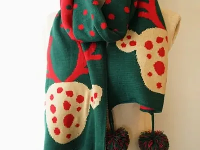 Grüner Strickschal mit Rentier-Allover-Muster und roten Punkten auf einer Schaufensterpuppe, mit dekorativen Pompom-Fransen an den Enden