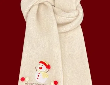 Weicher cremefarbener Strickschal mit aufgesticktem Schneemann-Motiv und roten Bommeln als Dekoration, auf dunkelrotem Hintergrund zusammengelegt