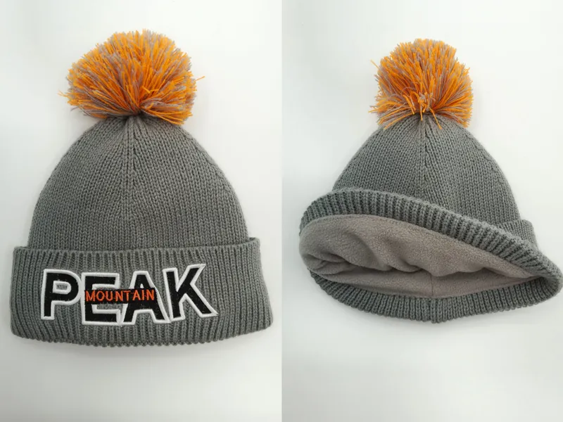 Graue Grobstrick-Wintermütze mit orangefarbenem Bommel, gesticktem Buchstaben-Patch und weichem Fleece-Innenfutter in Vorder- und Innenansicht