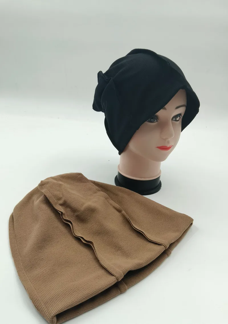 Lockere schwarze Strickmütze als Hijab-Unterziehmütze auf Modellkopf, daneben eine beigefarbene strukturierte Innenkappe auf weißem Untergrund