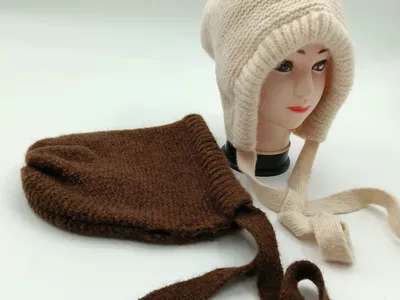 Cremefarbene Grobstrickmütze mit Ohrschutzklappen und geflochtenen Bändeln auf Hutständer, daneben braune Strickmütze mit langen Bindeschnüren auf weißem Hintergrund