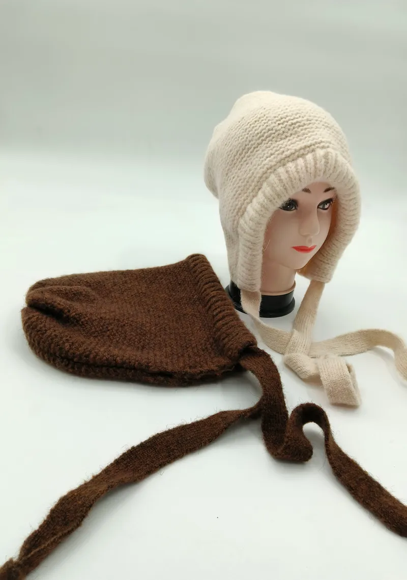 Cremefarbene Grobstrickmütze mit Ohrschutzklappen und geflochtenen Bändeln auf Hutständer, daneben braune Strickmütze mit langen Bindeschnüren auf weißem Hintergrund