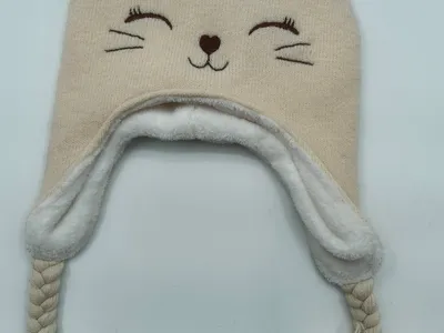 Cremefarbene gestrickte Wintermütze in Katzendesign mit Ohrklappen, geflochtenen Bindeschnüren, Pompons, weichem Fleecefutter und bestickten Katzengesichtsdetails