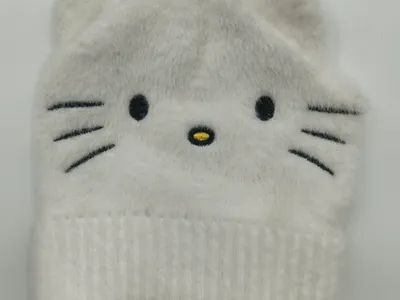 Weiße flauschige Strickmütze mit aufgesticktem Katzengesicht, Schnurrhärchen und kleinen spitzen Ohren, flach auf weißem Untergrund fotografiert