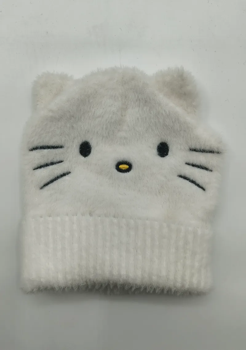 Weiße flauschige Strickmütze mit aufgesticktem Katzengesicht, Schnurrhärchen und kleinen spitzen Ohren, flach auf weißem Untergrund fotografiert