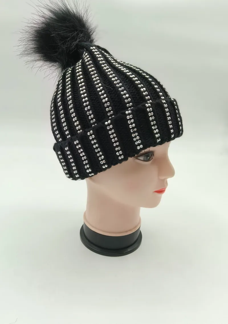 Schwarze Rippenstrickmütze mit silbernen Strassstreifen und schwarzem Kunstfell-Pompon auf Modellkopf präsentiert