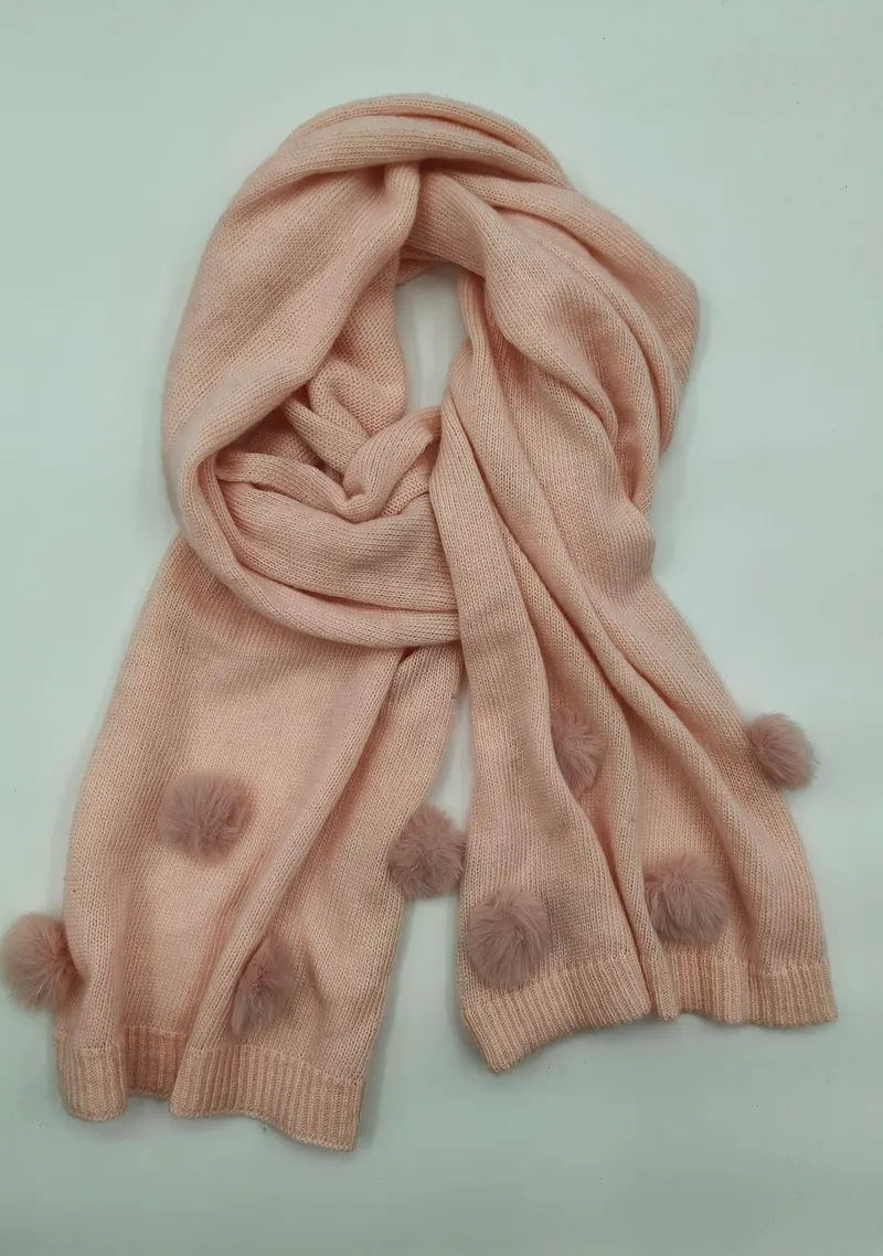 Hellrosa handgestrickter Winterschal mit dekorativen Wollbommeln als Randverzierung, locker aufgerollt vor weißem Hintergrund