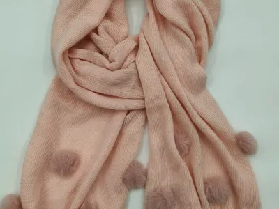 Hellrosa handgestrickter Winterschal mit dekorativen Wollbommeln als Randverzierung, locker aufgerollt vor weißem Hintergrund