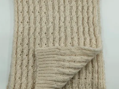 Gefaltete cremeweiße Strickdecke mit Reliefmuster aus Zopf- und Rippenstruktur auf weißem Untergrund