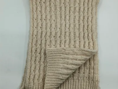 Gefaltete cremebeige Grobstrickdecke mit charakteristischem Zopf- und Rippenrelief auf weißem Untergrund