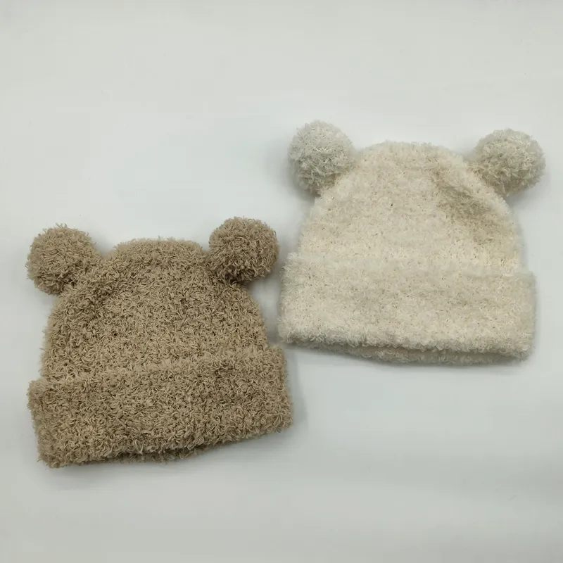 Zwei flauschige Babystrickmützen mit Bärenohren und Pompons in Beige und Creme auf weißem Hintergrund