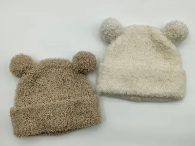 Zwei flauschige Babystrickmützen mit Bärenohren und Pompons in Beige und Creme auf weißem Hintergrund