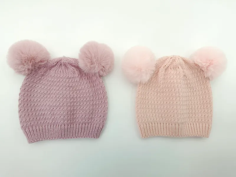 Zwei gestrickte Winterbeanies mit je zwei Kunstfell-Pompons in den Farben Flieder und Hellrosa, auf weißem Hintergrund fotografiert