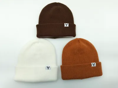 Drei gefaltete Rippenstrick-Beanies mit Umschlagbund in Dunkelbraun, Cremeweiß und Karamellorange, jede mit kleinem aufgenähtem Markenpatch