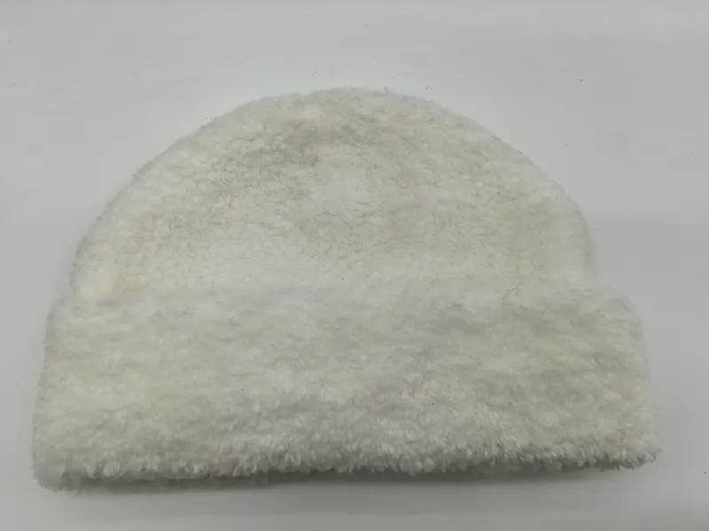 Cremefarbene Plüschmütze aus Sherpa-Fleece mit lockiger Teddystruktur, auf hellem Hintergrund liegend – flauschiges Winteraccessoire