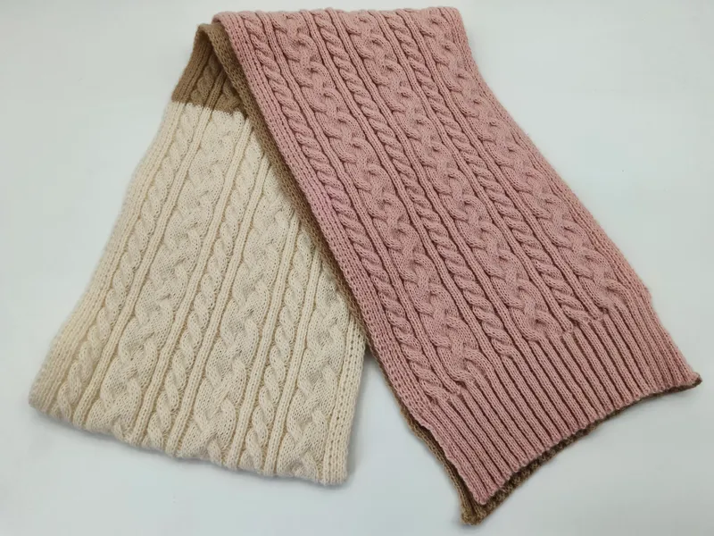 Zwei Zopfstrick-Schals zusammengefaltet, einer in Creme und einer in Altrosa, mit beigen Rippenrändern auf weißem Hintergrund