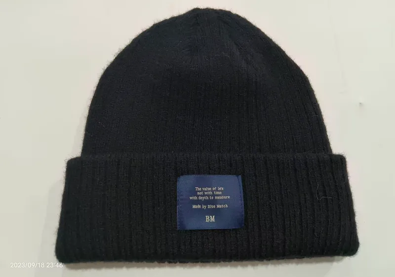 Schwarze Rippenstrickmütze mit gefaltetem Rand und blau-gewebtem Patch-Etikett auf hellem Untergrund