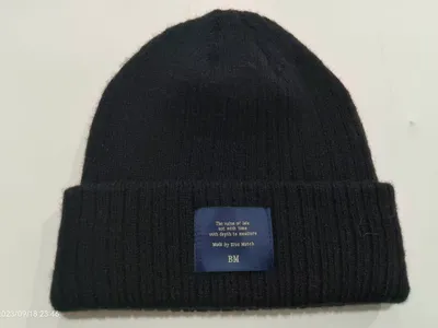 Schwarze Rippenstrickmütze mit gefaltetem Rand und blau-gewebtem Patch-Etikett auf hellem Untergrund