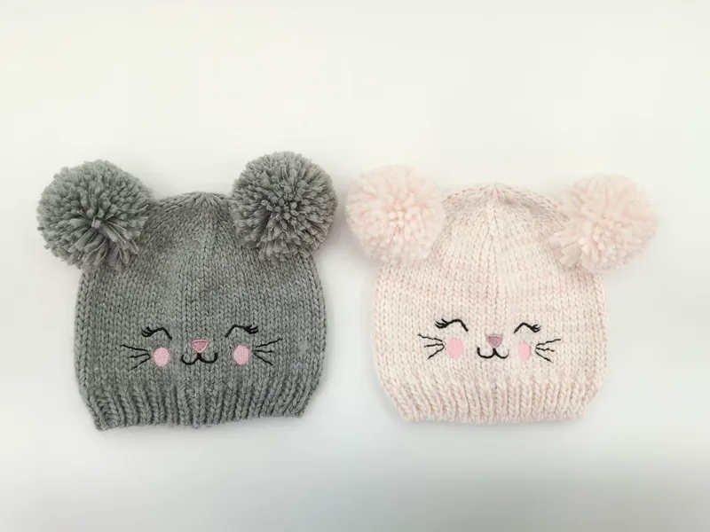 Zwei handgestrickte Babymützen mit flauschigen Bommel-Ohren und aufgestickten Katzengesichtern, eine in Grau und eine in Hellrosa, auf weißem Hintergrund