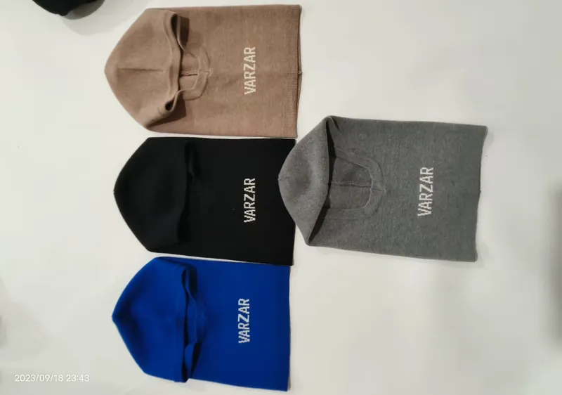 Vier gestrickte Sturmhauben in Beige, Schwarz, Grau und Blau flach auf weißer Fläche, mit besticktem Markenlogo