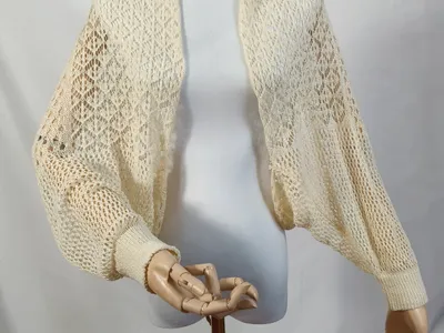 Elfenbeinfarbenes Häkel-Bolero mit weiten Bischofärmeln und gerippten Bündchen auf einer Schneiderpuppe in Frontansicht