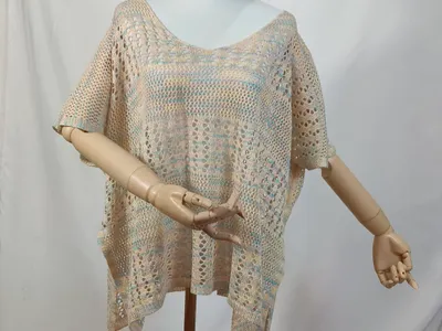 Beigefarbenes Multicolor-Häkelponcho mit Kurzarm auf einer Schneiderpuppe mit Holzarmen, Pastellblau- und Rosatöne sichtbar