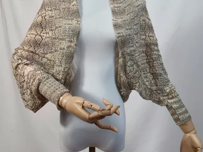 Offenes Spitzenstrick-Shrug in Beige-Grau-Mélange auf einer Schneiderpuppe mit Holzhänden, Fledermausärmel und Rippbündchen sichtbar