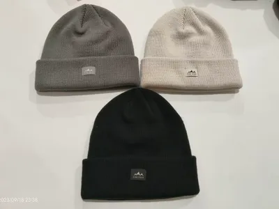 Drei gerippte Strickmützen mit Umschlagbund in Schwarz, Grau und Cremeweiß flach liegend, mit Berglogo-Patch
