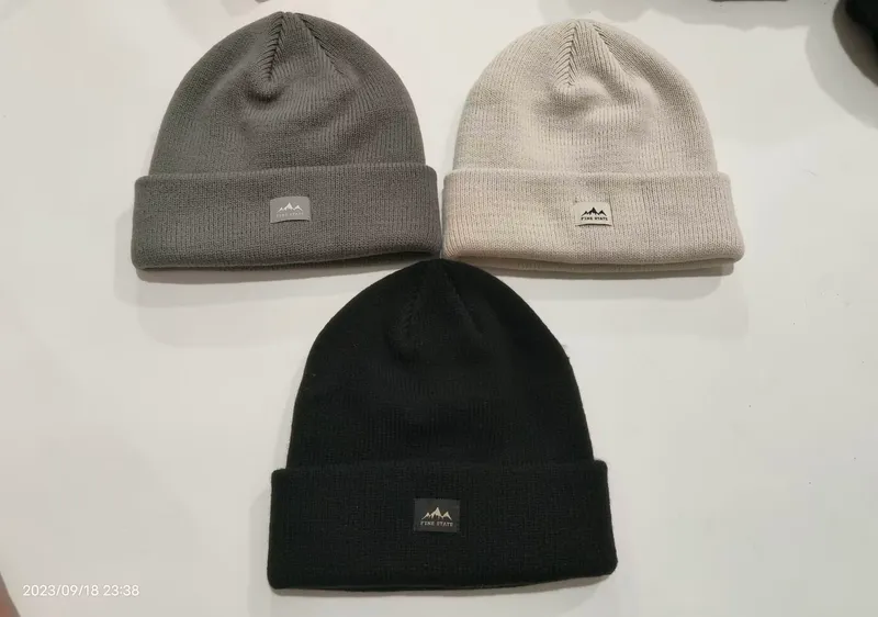 Drei gerippte Strickmützen mit Umschlagbund in Schwarz, Grau und Cremeweiß flach liegend, mit Berglogo-Patch